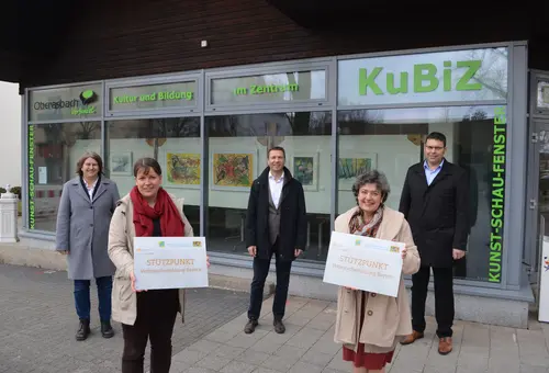 Fünf Personen stehen vor dem Kunstschaufenster Oberasbch. Zwei Frauen im Vordergrund präsentieren das Schild Stützpunk Verbraucherbildung Bayern