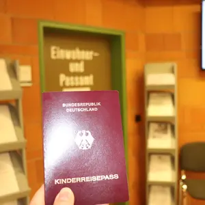 Eine Hand hält einen roten Kinderreisepass der Bundesrepublik. Im Hintergrund ist die Tür des Einwohner- und Passamts zu sehn. 
