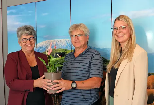 Eine Frau überreicht einem Mann eine Topfpflanze, rechts daneben steht eine junge Frau