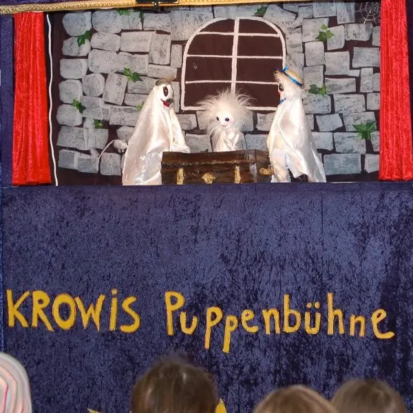 Blick auf eine Puppenbühne. Vor einer groben Steinmauer mit einem runden Fenster sitzen drei Gespenster an einem dunkelbraunen Tisch. 