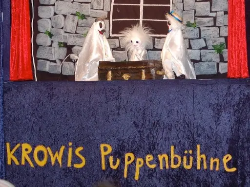 Blick auf eine Puppenbühne. Vor einer groben Steinmauer mit einem runden Fenster sitzen drei Gespenster an einem dunkelbraunen Tisch. 