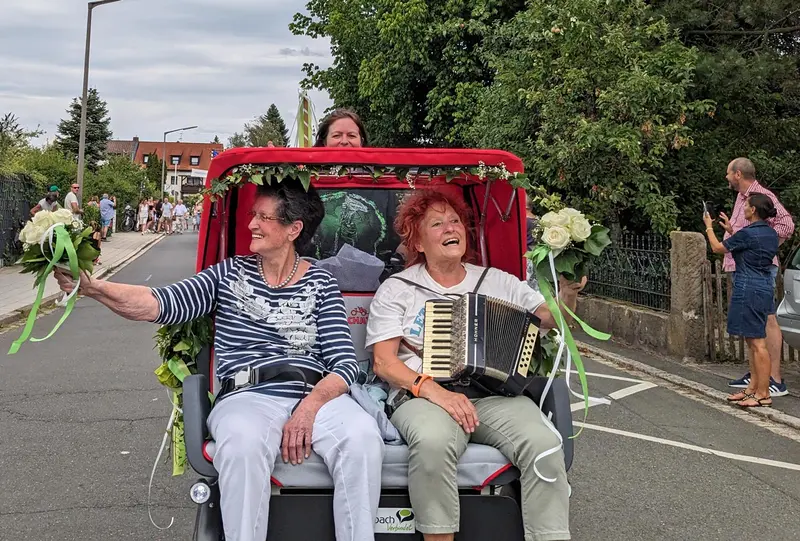 zwei Frauen in einer E-Rikscha (frontal); die linke grüßt jmd außerhalb des Bildes; die rechte spielt Akkordeon. Halb hinter dem Verdeck sieht man die Fahrerin 